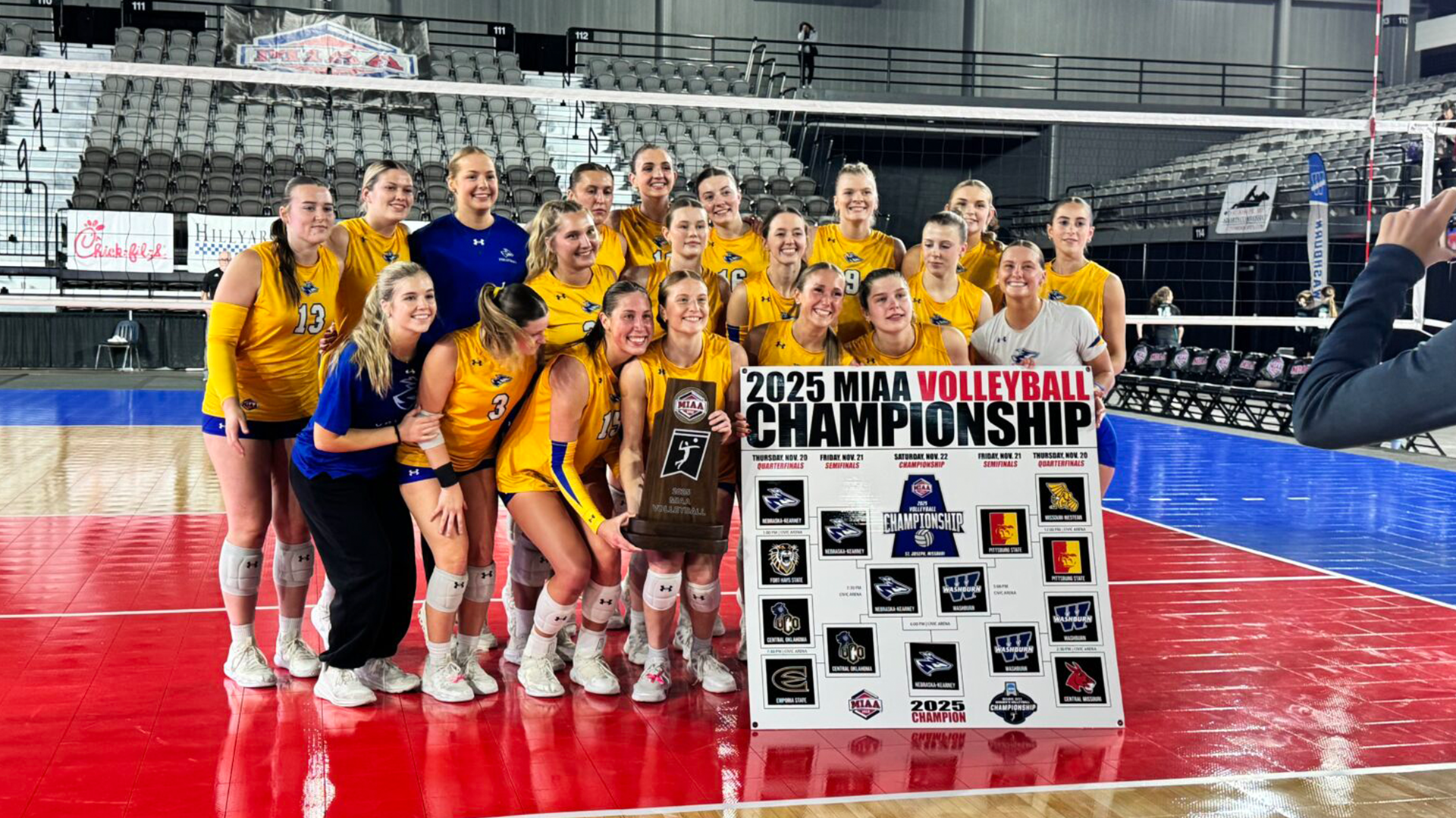 Nebraska Kearney volleyball players celebrate with the 2025 MIAA Volleyball Championship trophy and tournament bracket at St. Joseph Civic Arena.