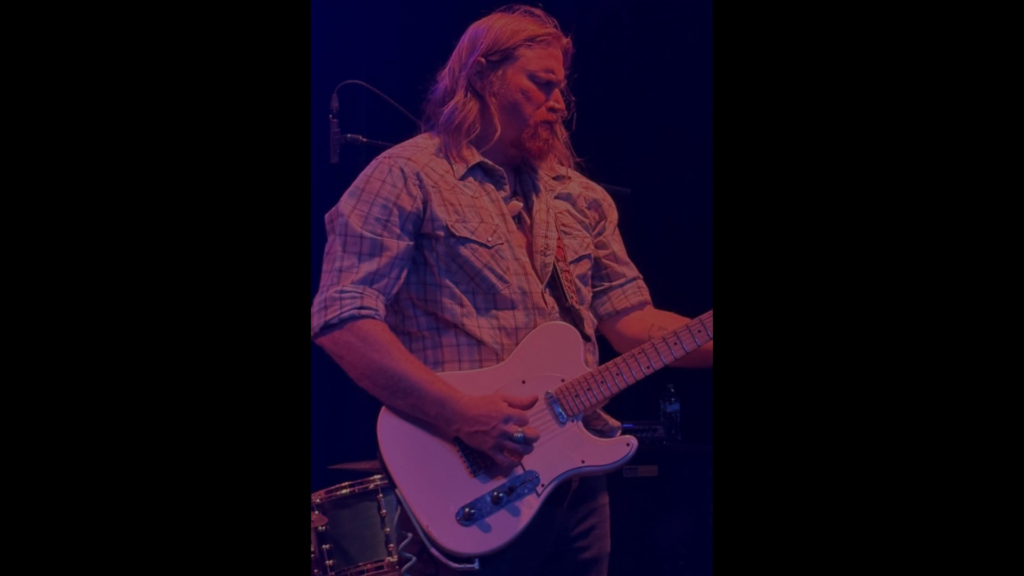 Kris Lager performs on electric guitar under purple stage lighting during a live concert.
